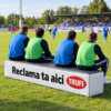 Trei adulti stau pe o banca de rezerve din burete personalizata, pe marginea unui teren de fotbal